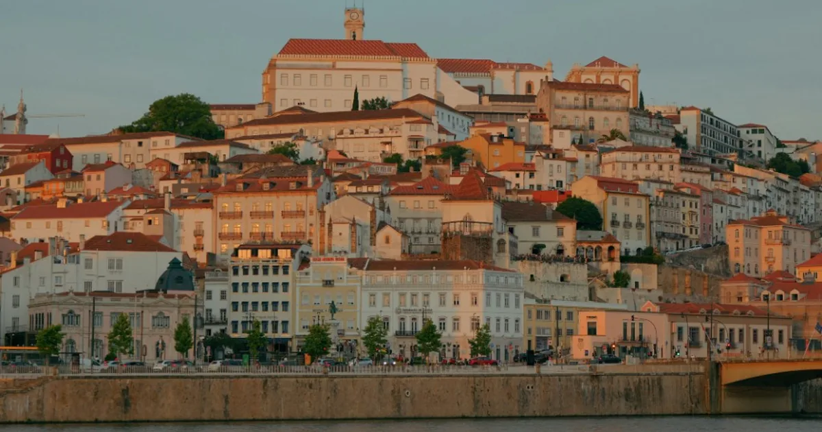 Hillside of cream buildings with terracotta roofs rising to a white clock-tower building, above a river and a bridge
