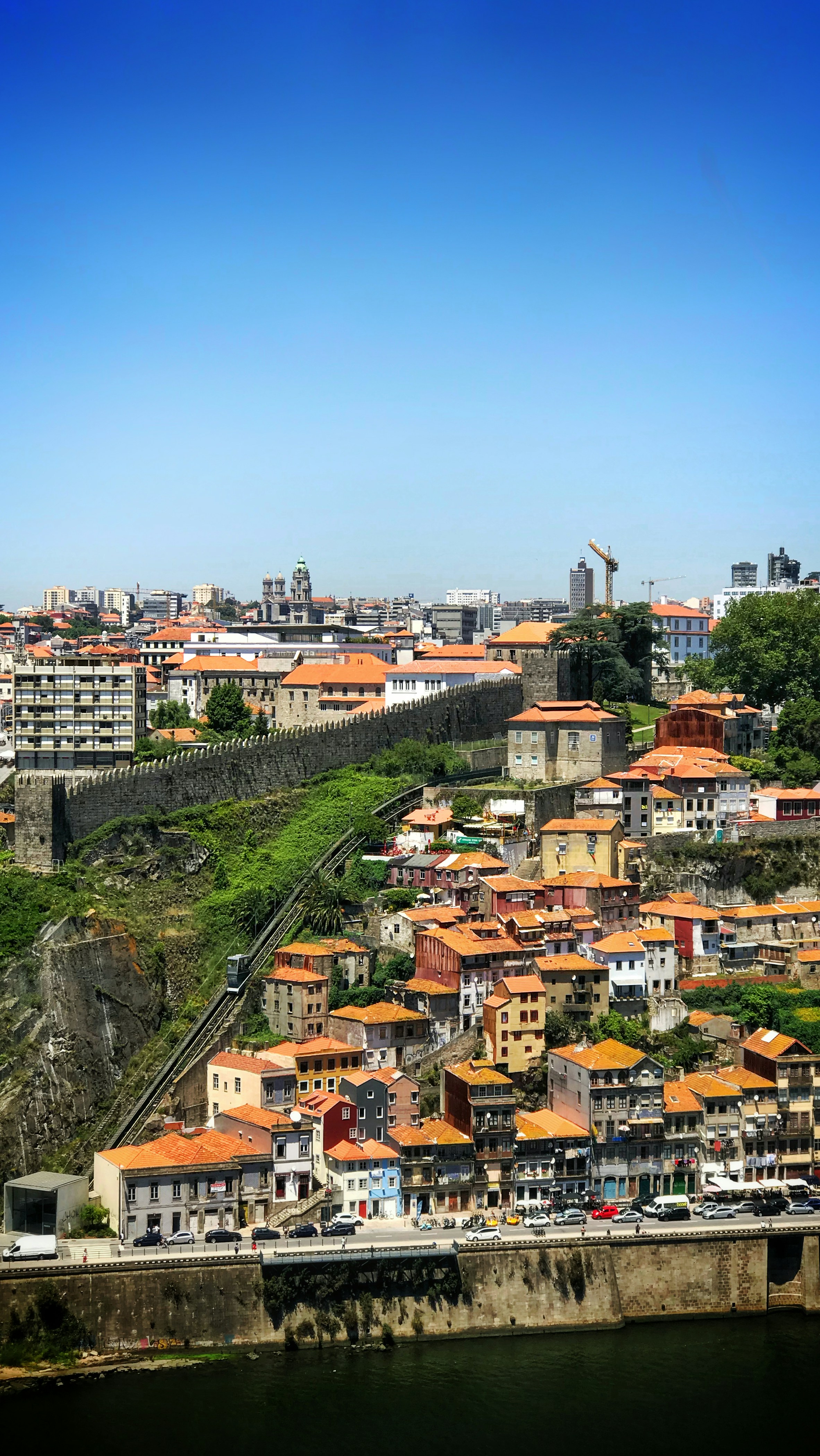 Stone city wall above orange-roofed houses and the Douro waterfront in Porto, under a clear blue sky