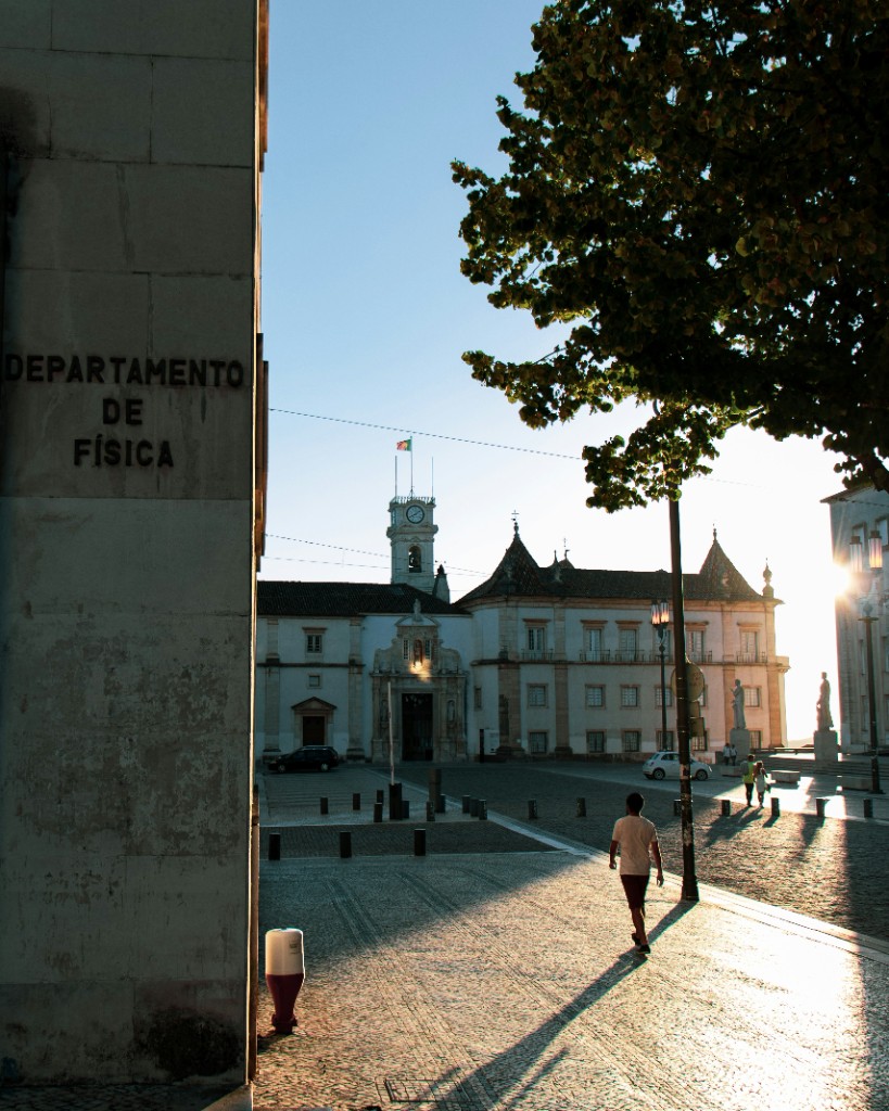 A person walks across a sunlit cobblestone plaza toward a clock-tower building, beside a wall labeled Departamento de Física