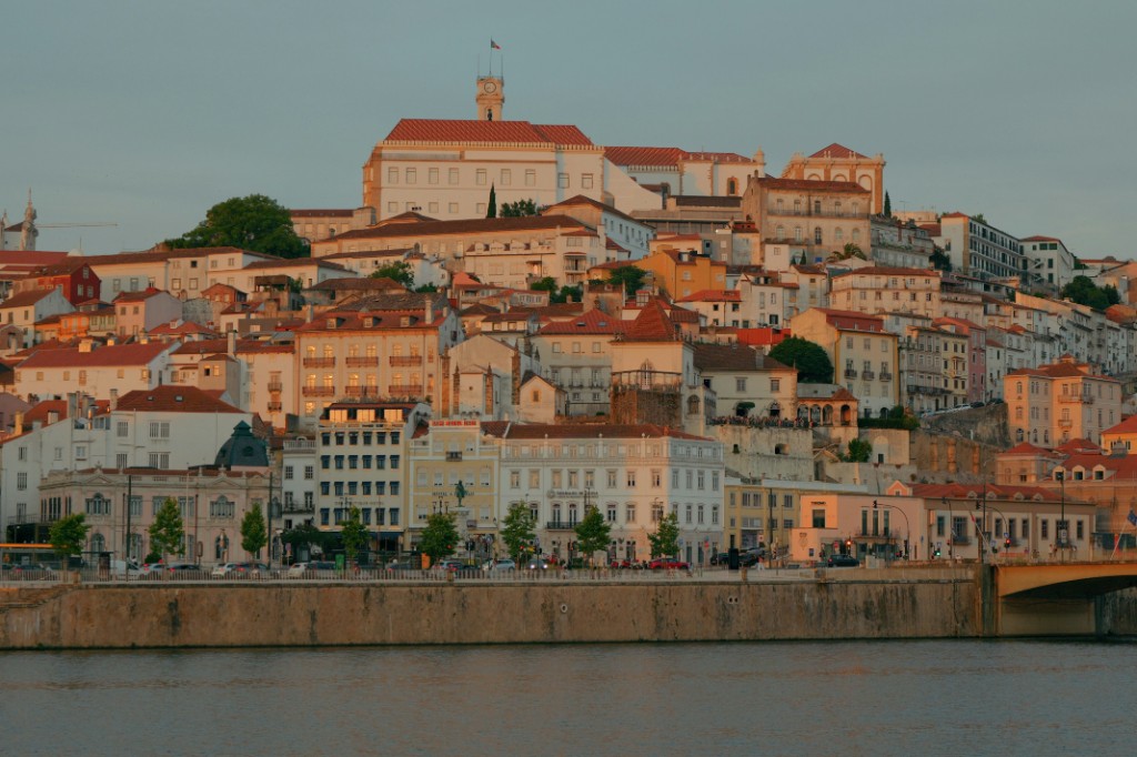 Hillside of cream buildings with terracotta roofs rising to a white clock-tower building, above a river and a bridge