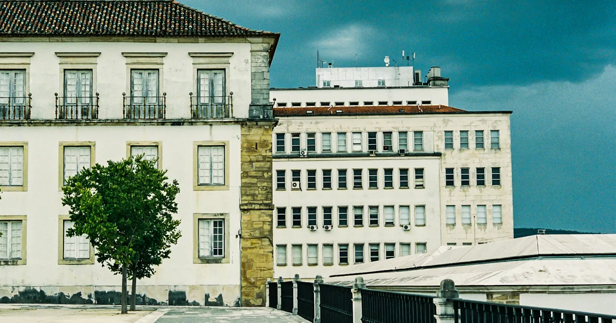 White historic facade beside a later office block, with a tree, paved walk, and iron railing