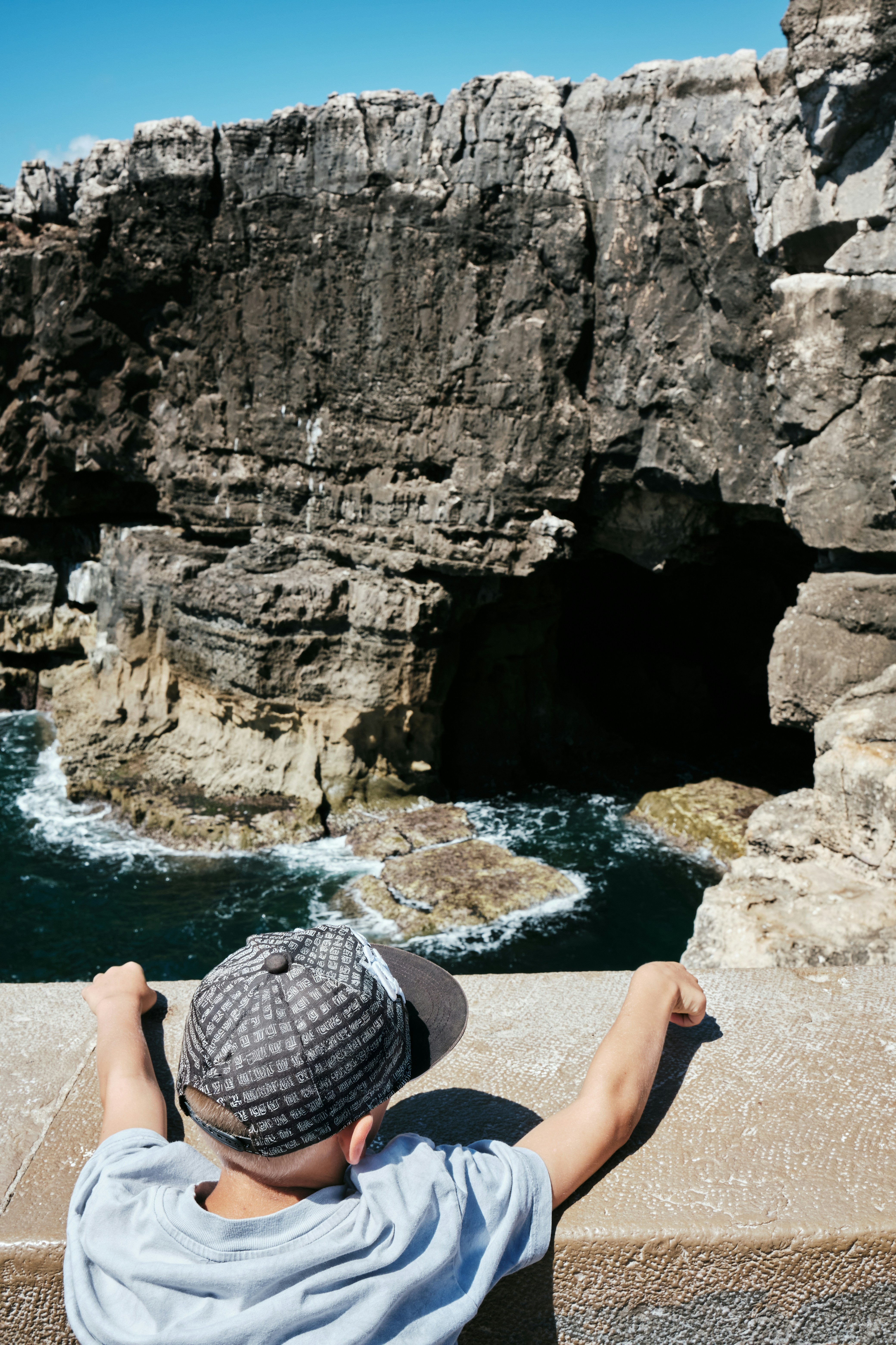 Child in a patterned cap leaning on a stone wall, looking down at a sea cave in dark cliffs