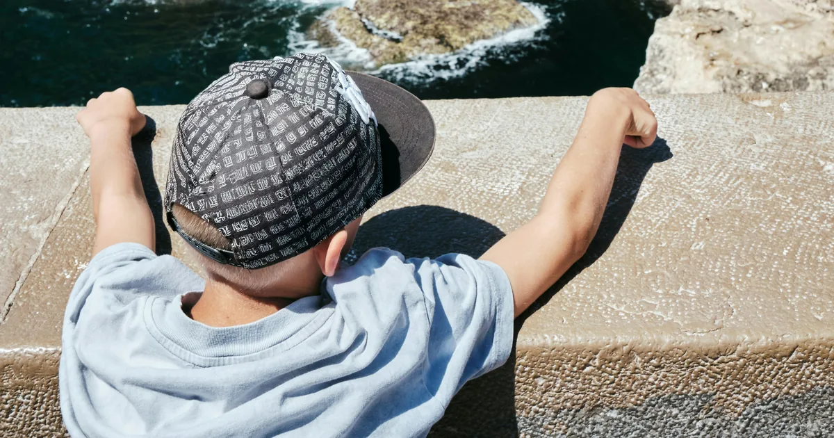 Child in a patterned cap leaning on a stone wall, looking down at dark sea cliffs