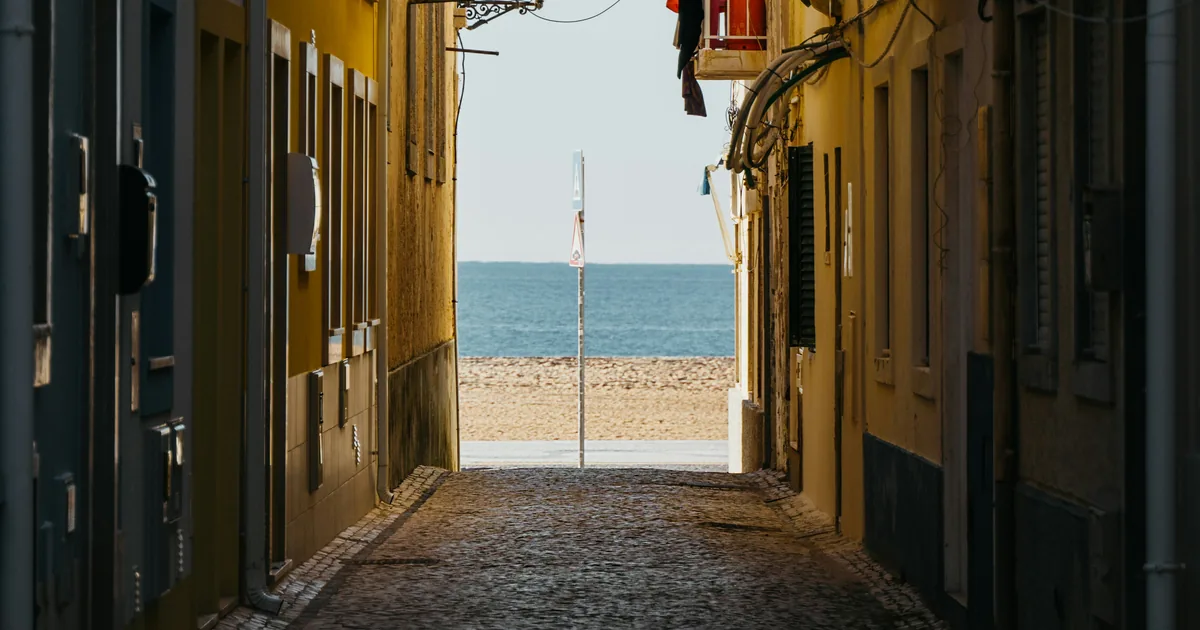Narrow cobbled lane between yellow and cream houses that opens onto a beach and the sea