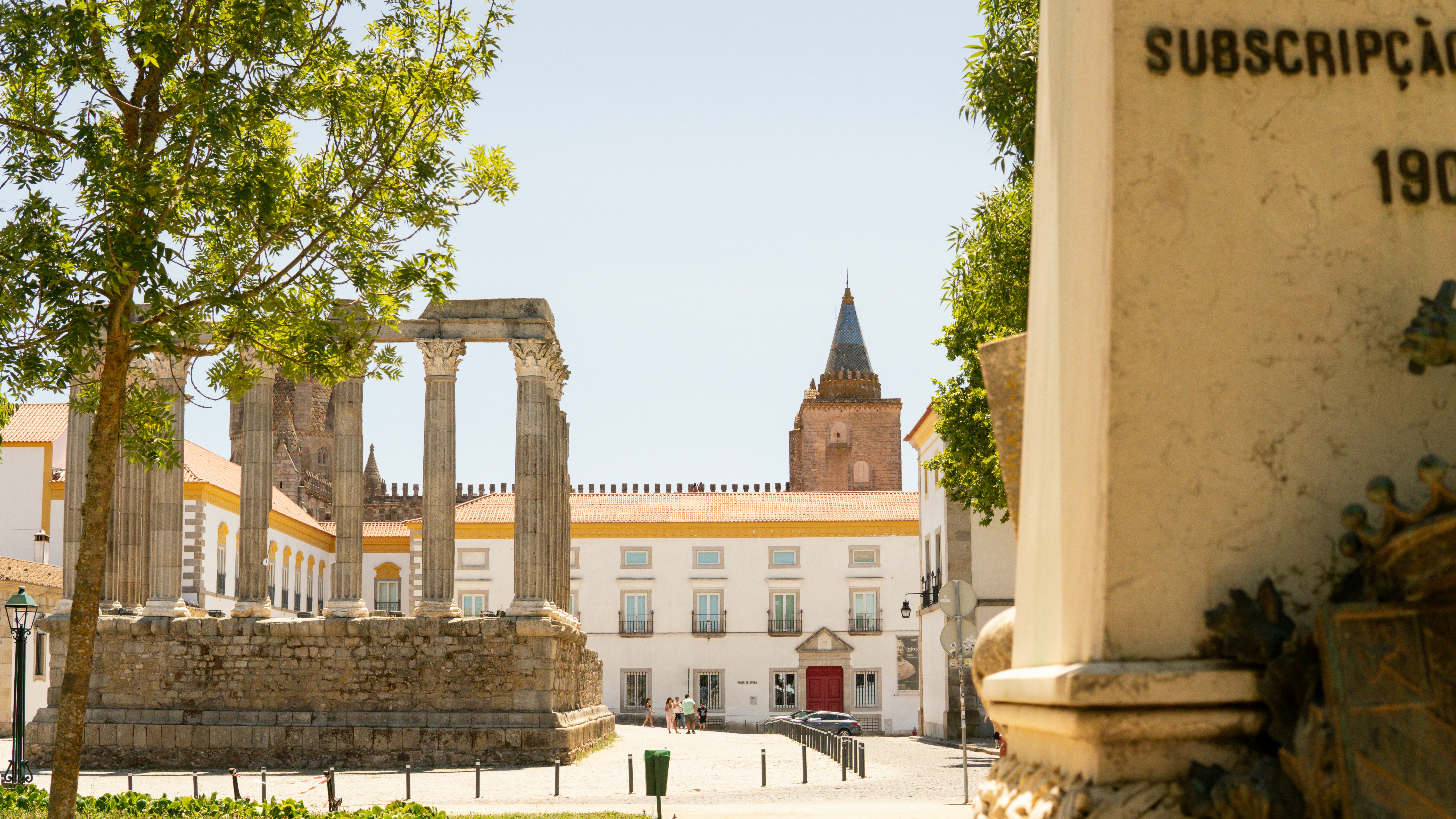 Roman temple columns in an Évora square facing a white palace and church tower, with a carved Subscrição inscription in the foreground
