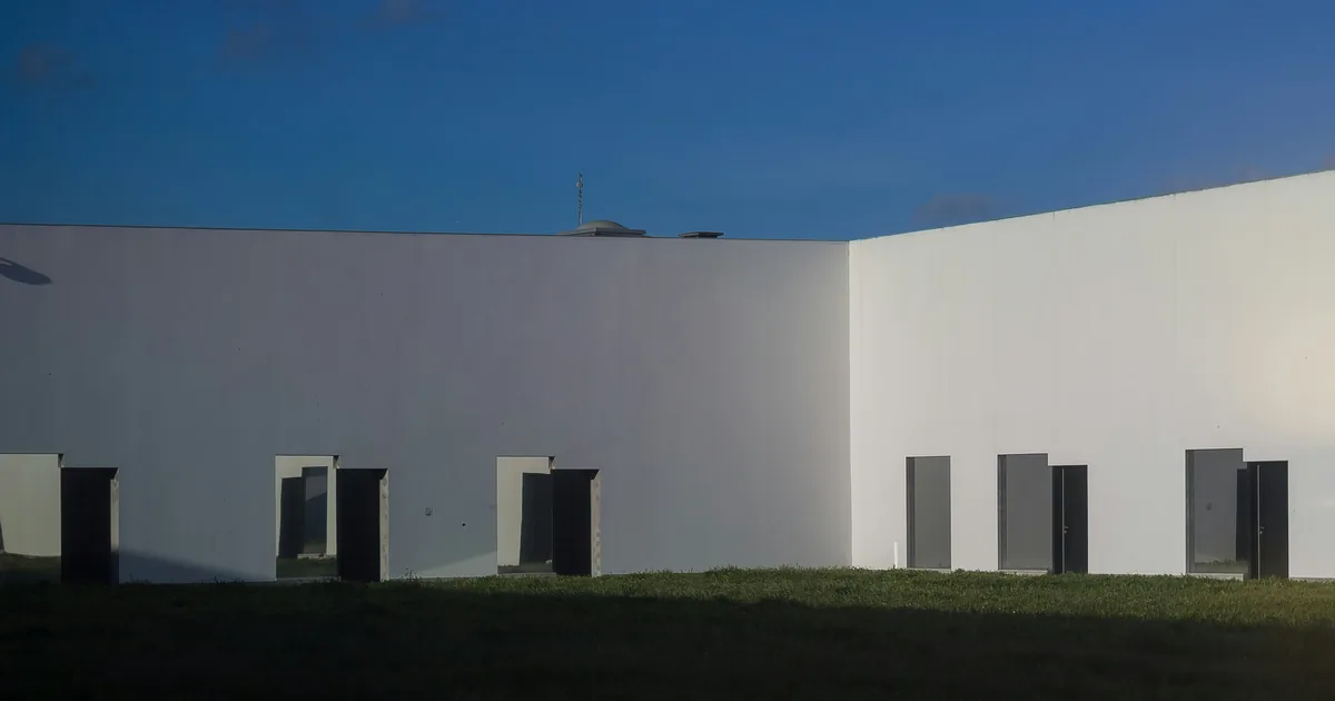 White courtyard walls with dark doorways opening onto grass under a blue sky