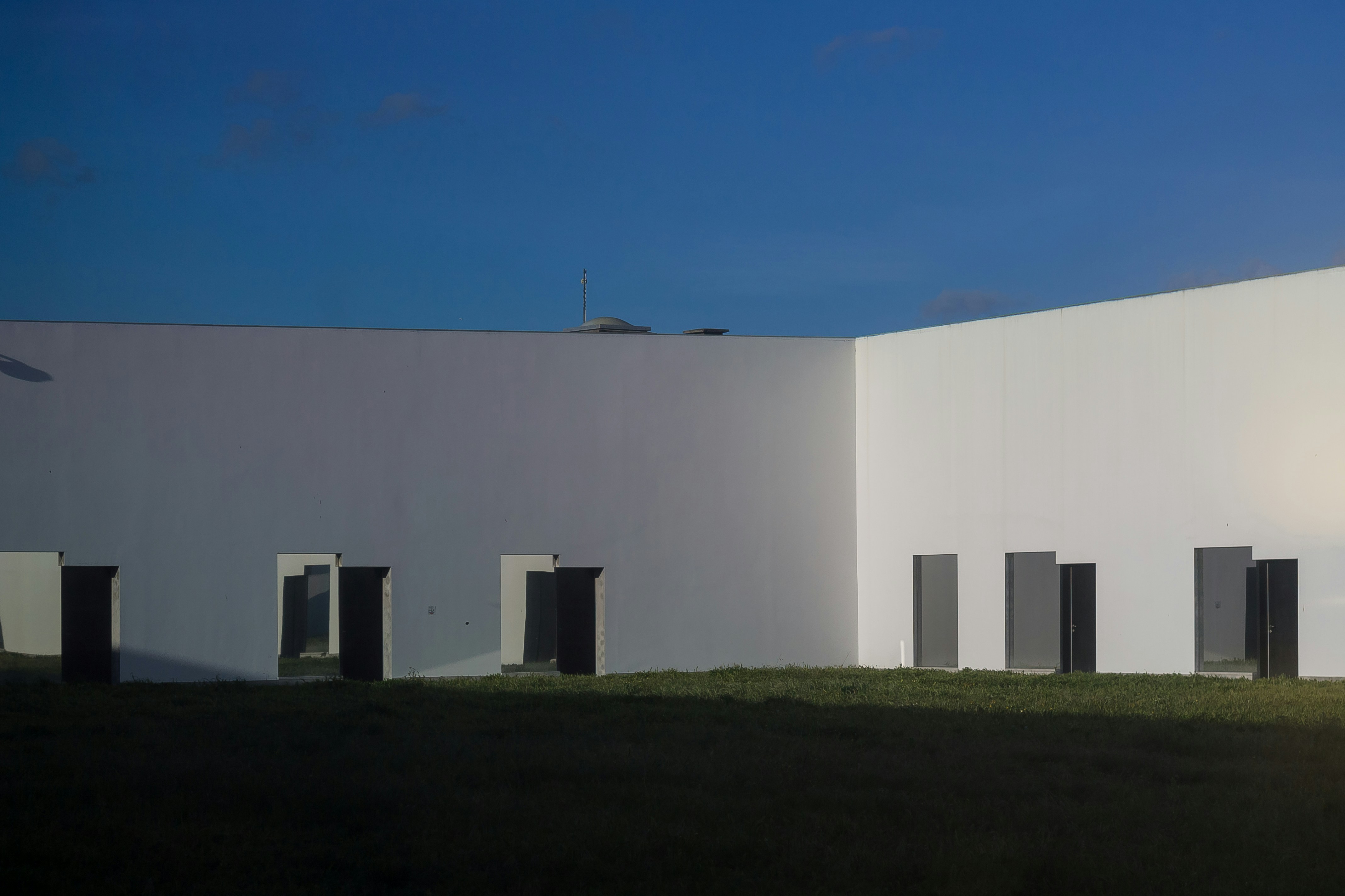 White courtyard walls in cooler light, with dark doorways opening onto grass under a blue sky