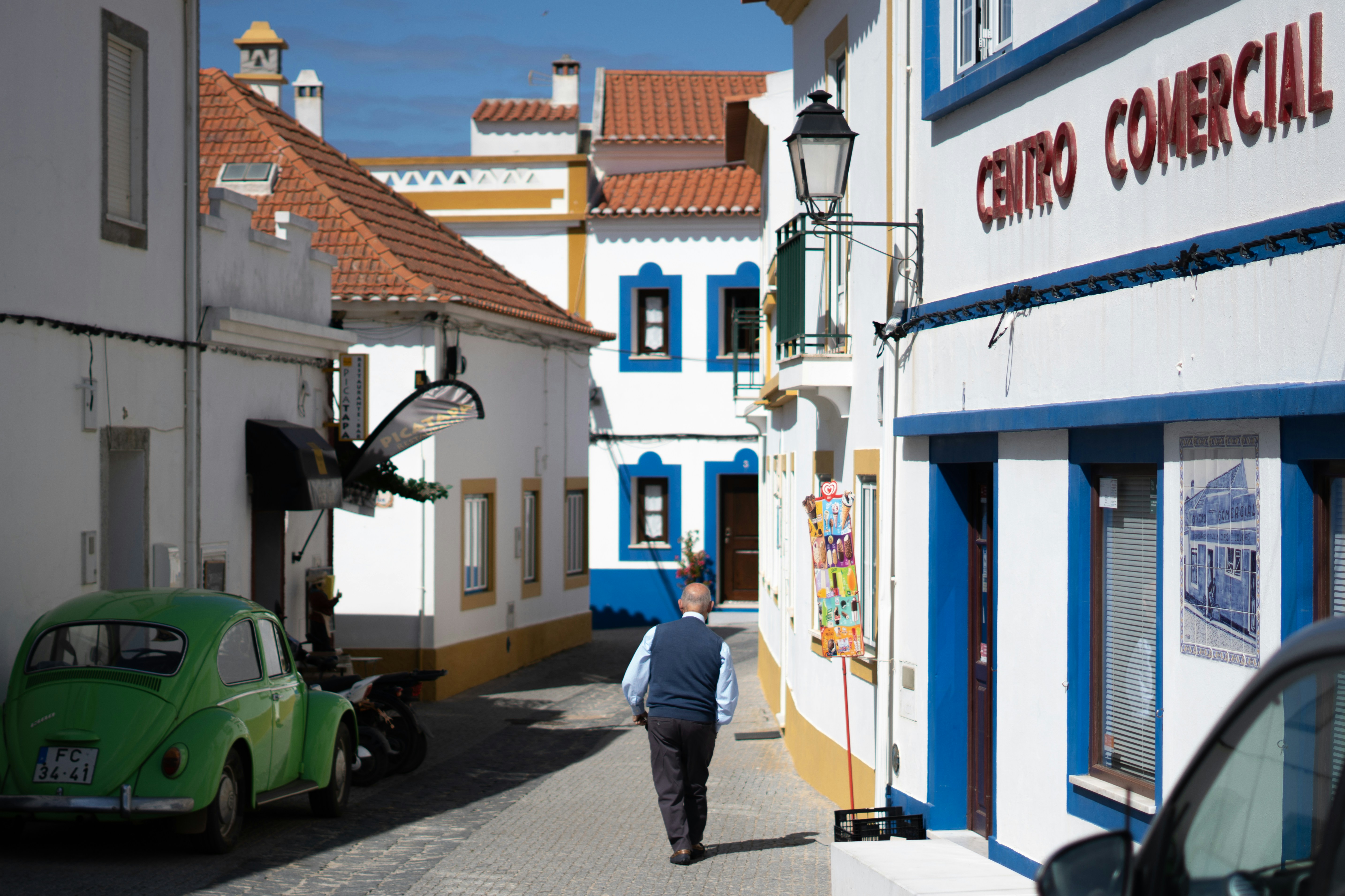 Man walking away down a cobbled lane past a green Volkswagen Beetle and a Centro Comercial sign