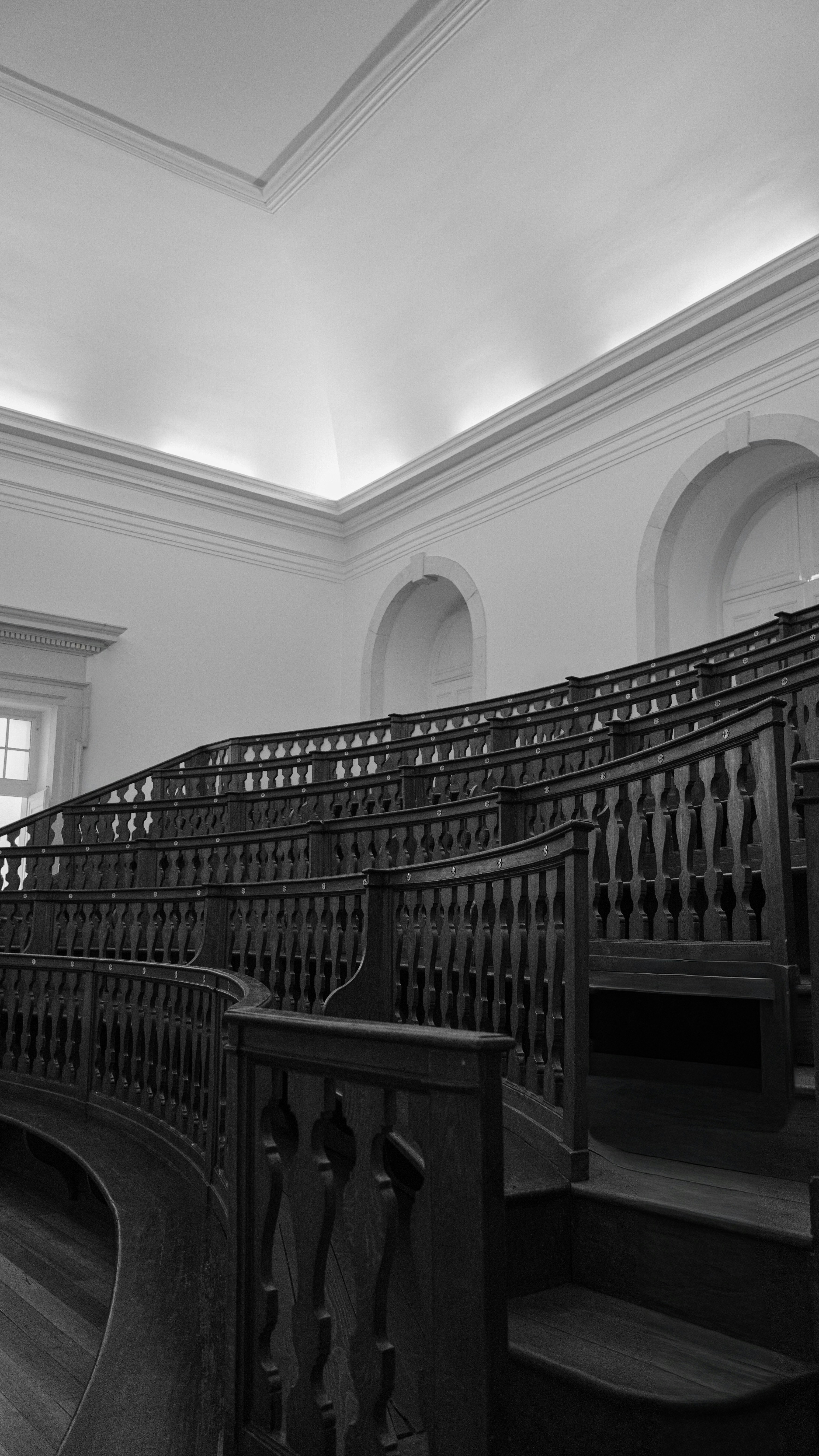 Empty dark wooden benches rising in a curved lecture hall with white walls and arched windows