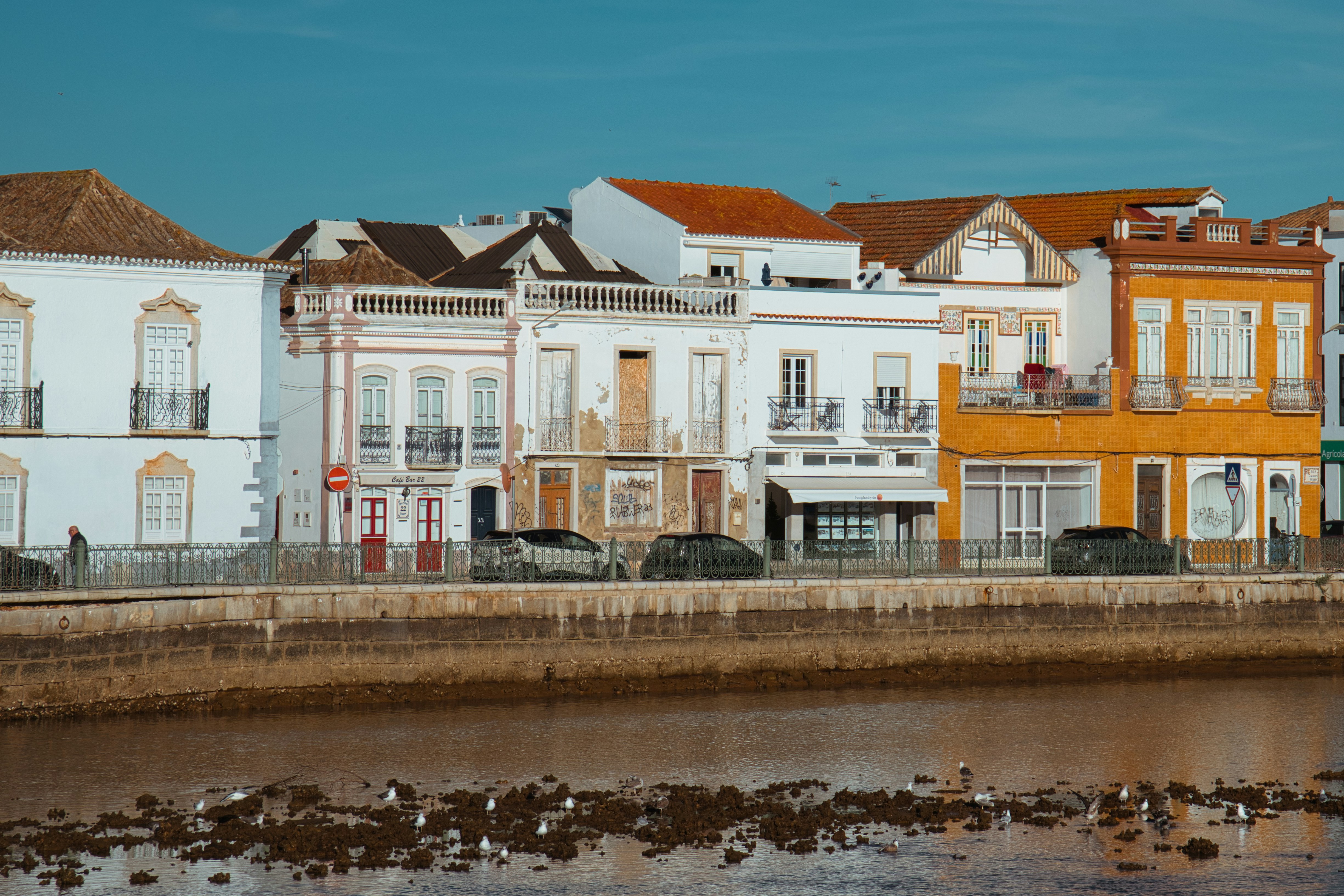 Row of white and ochre houses along a tidal mudflat, with parked cars and wading birds