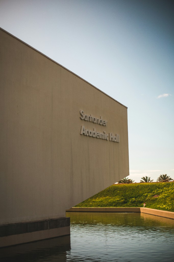 Santander Academic Hall beside a reflecting pool, with lettering on the concrete facade and palms on the slope beyond