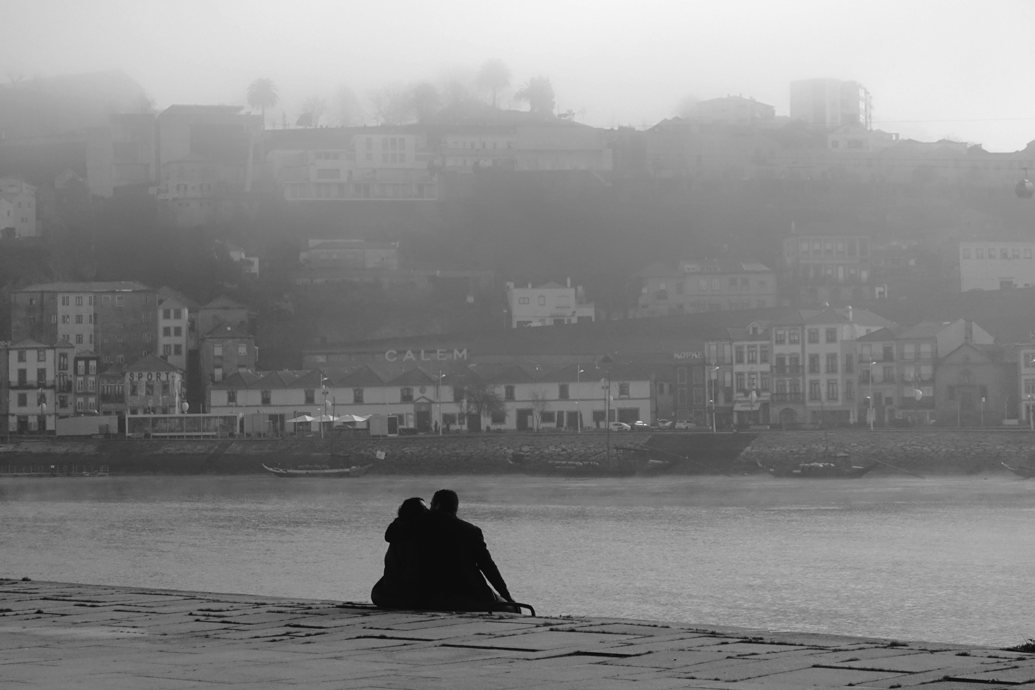 Couple sitting together on a riverside quay in mist, facing warehouses across the water