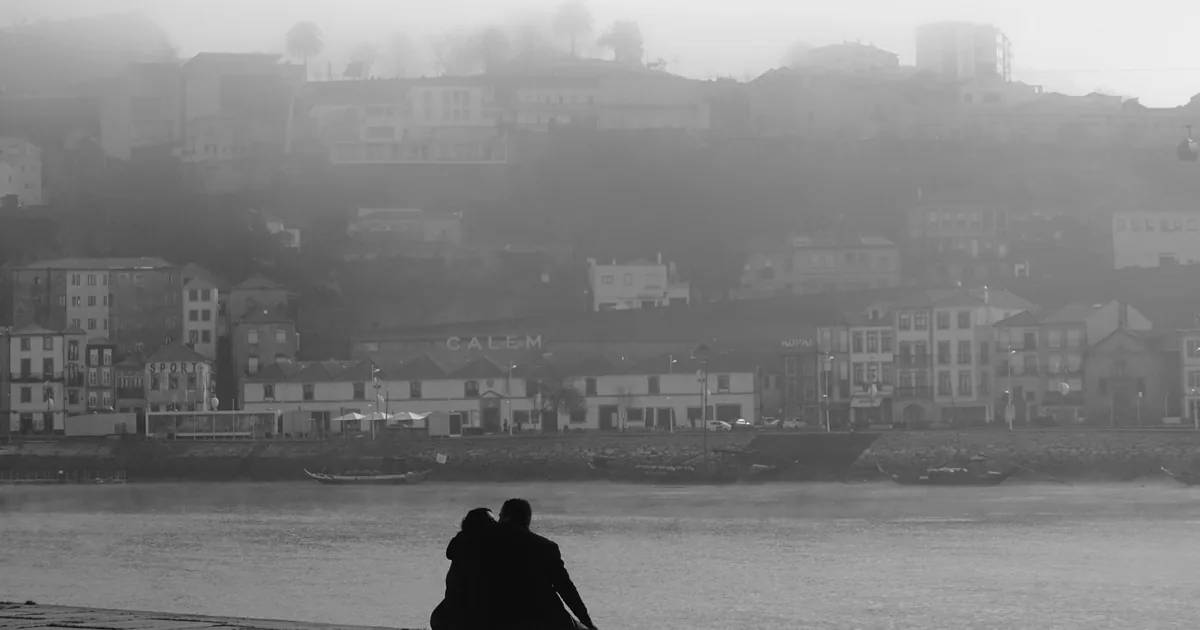 Couple sitting together on a riverside quay in mist, facing warehouses across the water