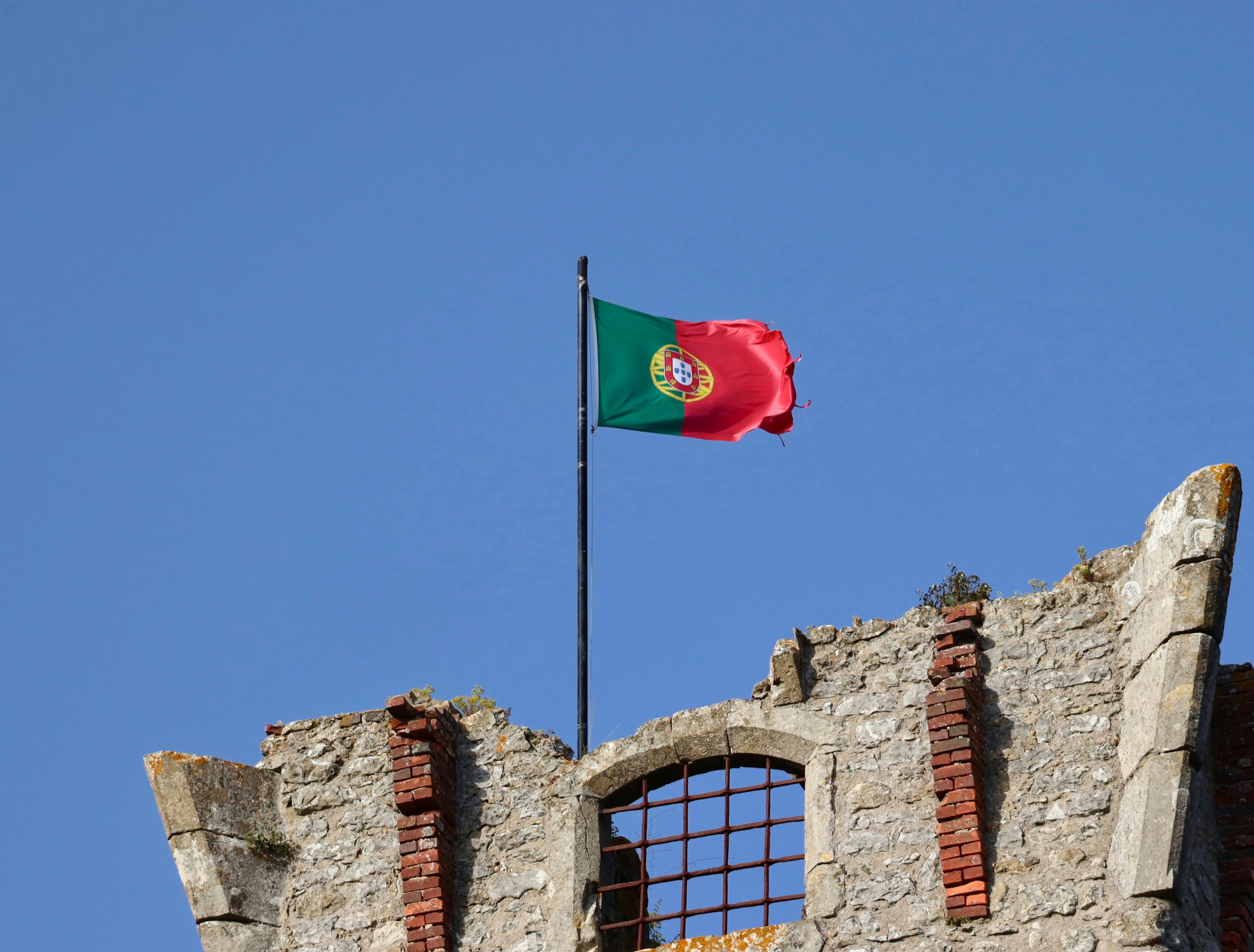 Portuguese flag on a black pole above a ruined stone wall with a barred window and brick repairs