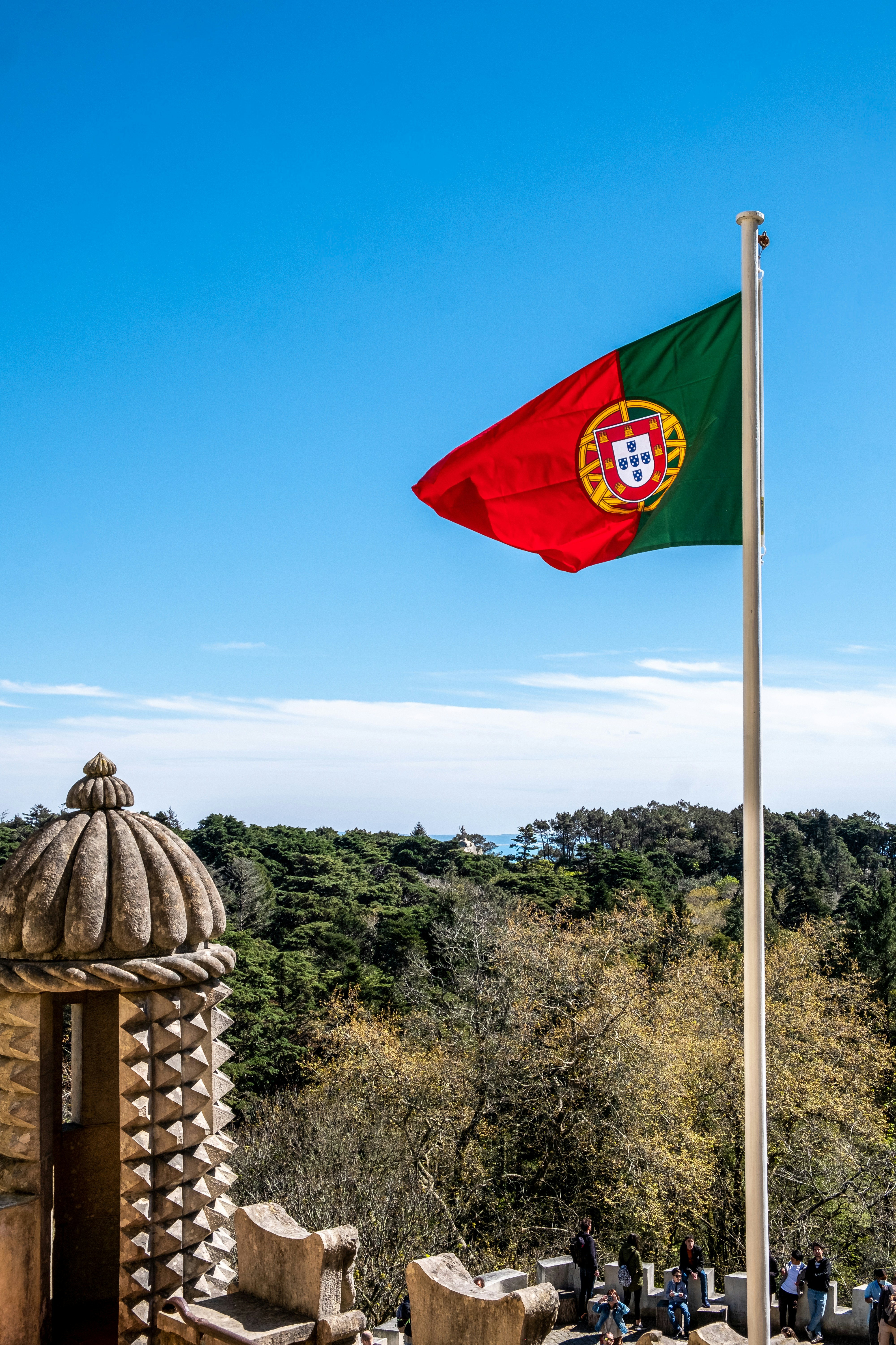 Portuguese flag on a white pole above pine woods, with a stone pineapple turret and visitors on a terrace
